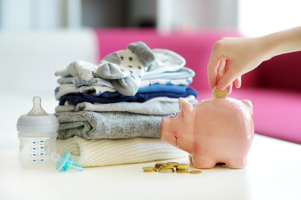 A hand putting a coin into a piggy bank next to a pile of folded baby clothes